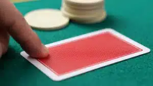深綠色百家樂桌面上，指尖按住一張蓋著的紅色牌背，後方是一疊籌碼
